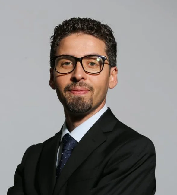 Un homme à lunettes et cheveux foncés, vêtu d’un complet noir, d’une chemise blanche et d’une cravate foncée à pois, debout sur un fond gris clair.