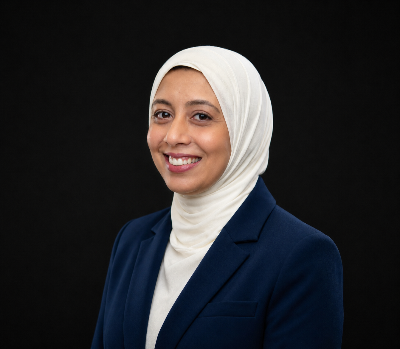Une femme portant un hijab blanc et un blazer bleu marine sourit sur fond noir.