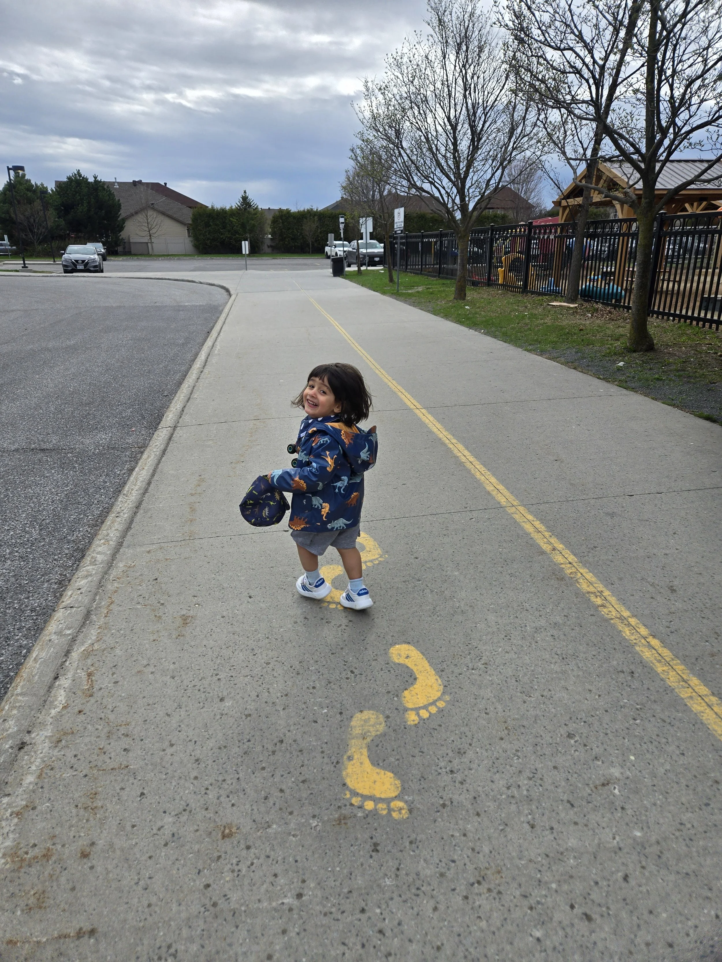 Un enfant souriant marchant sur un trottoir, portant un chapeau, avec des arbres et des voitures garées en arrière-plan, sous un ciel nuageux.