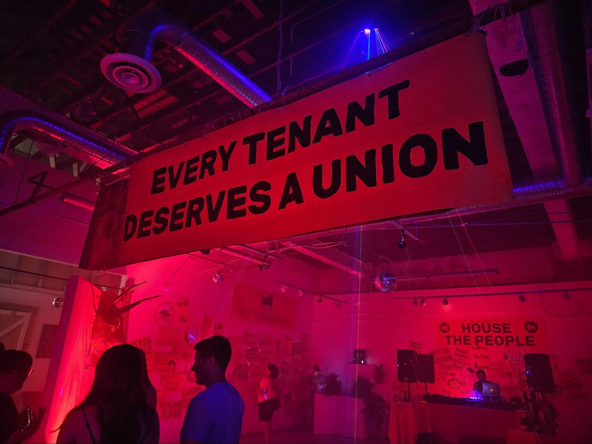 A large red banner with black text that says, "Every Tenant Deserves a Union" hanging from the ceiling of a dimly lit indoor space with pink and purple lighting. People silhouettes are visible below the banner.