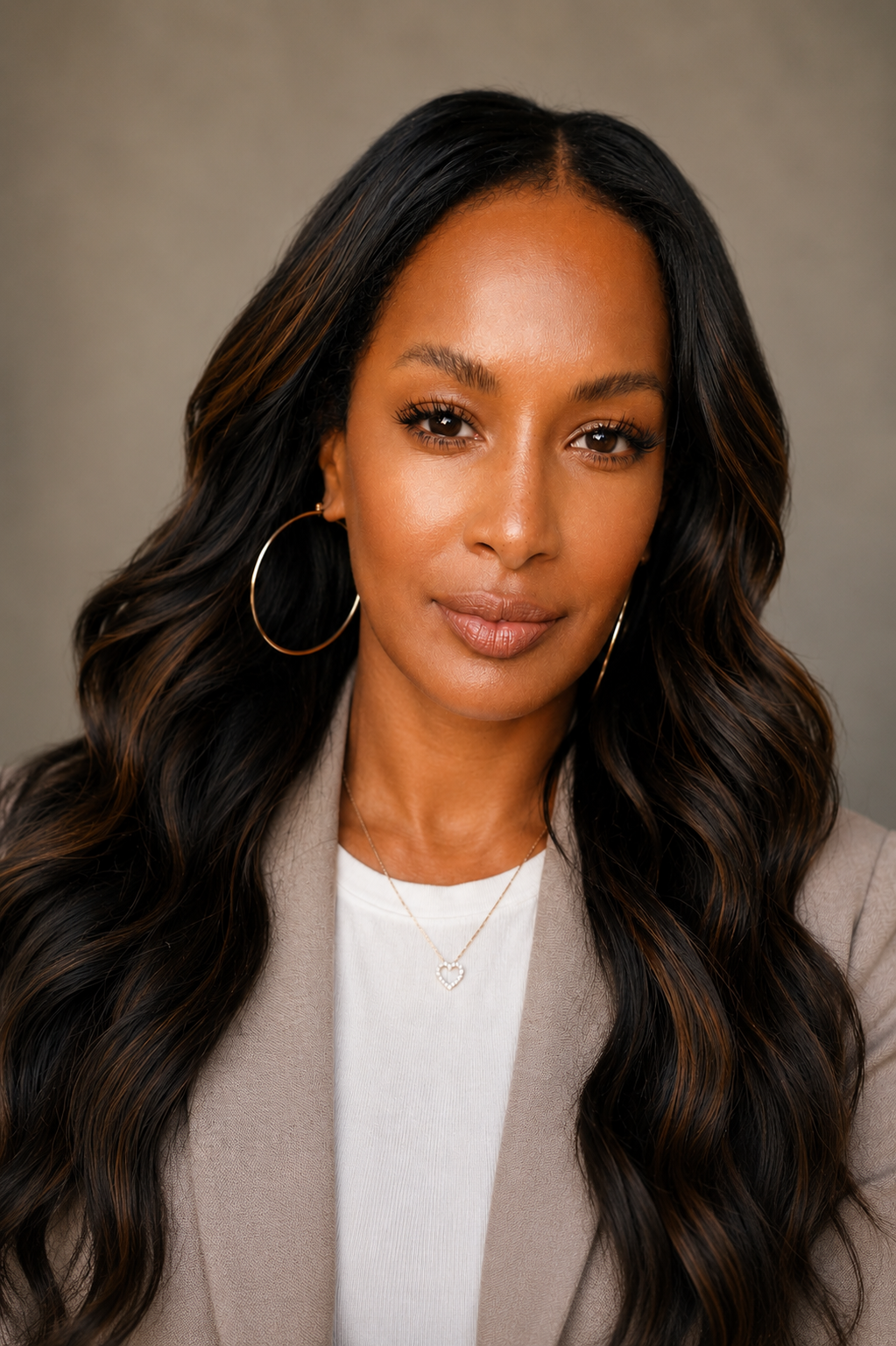 Close-up portrait of a woman with long, wavy black hair, hoop earrings, a necklace with a heart pendant, wearing a beige blazer and a white top, against a neutral background.
