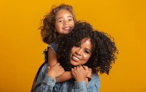 A woman and a young girl with curly hair smiling against a yellow background. The girl is on the woman's back with her arms around her neck.