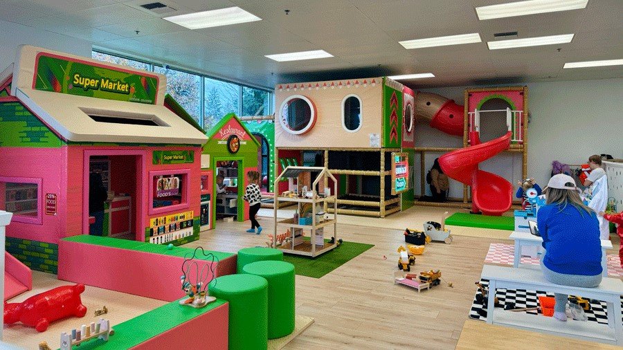Indoor children's play area designed like a colorful supermarket with play structures, a slide, and toys, with kids and adults playing.