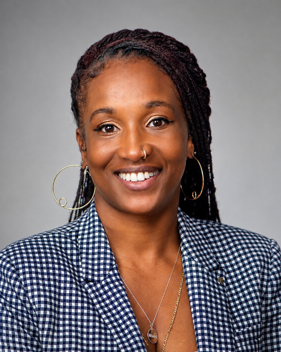 A woman with dark braided hair, hoop earrings, and a nose ring, smiling, wearing a checkered blazer and layered necklaces against a neutral grey background.