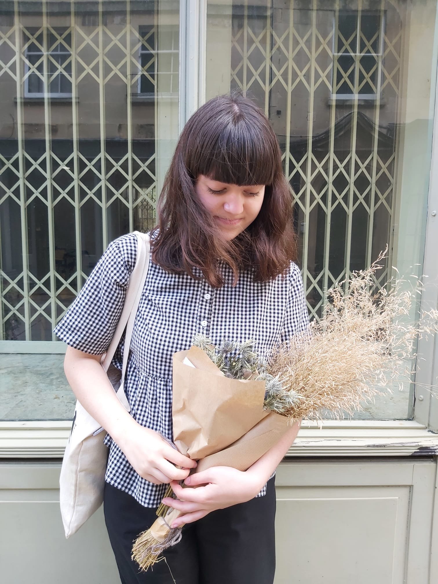 A young woman with long brown hair and bangs, wearing a black and white checkered shirt, holding a brown paper bag with a bouquet of dried flowers.