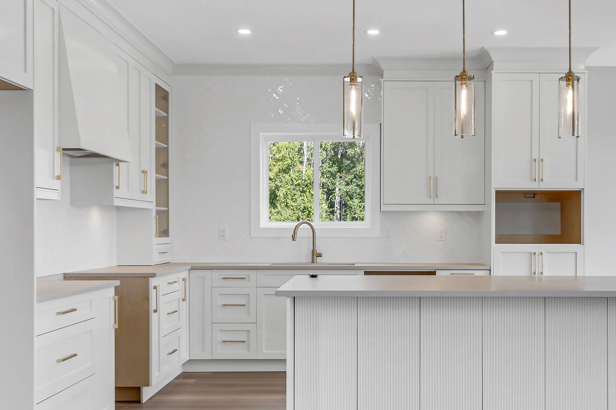 Modern white kitchen with gold hardware and pendant lights, a window overlooking greenery, and a counter with an integrated sink.
