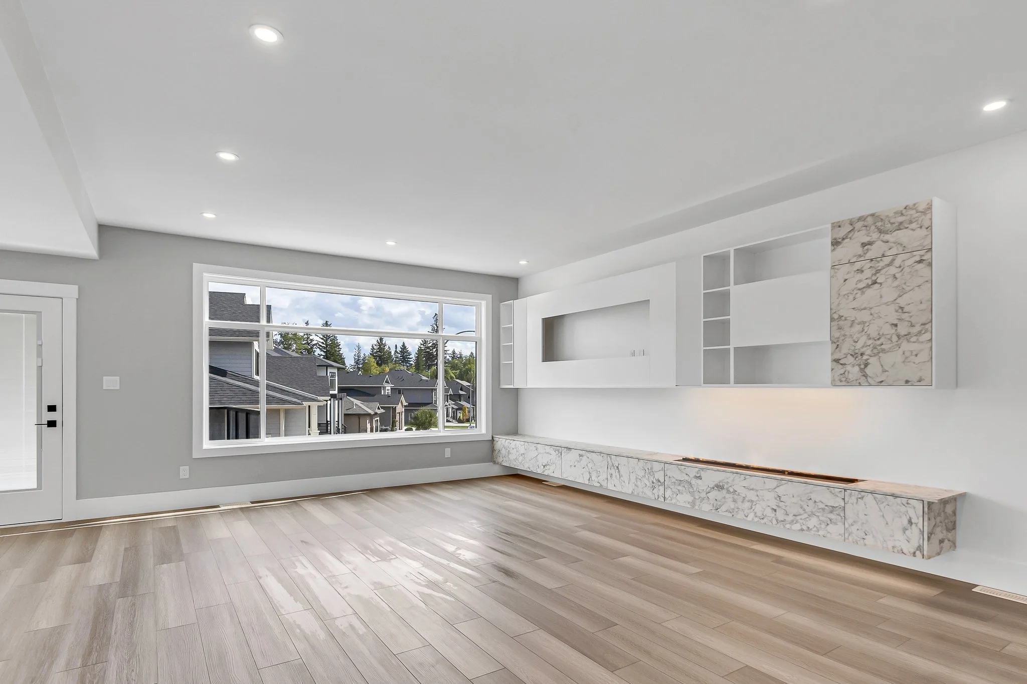 Empty living room with large window, light wood flooring, white walls, and built-in marble and white shelving.