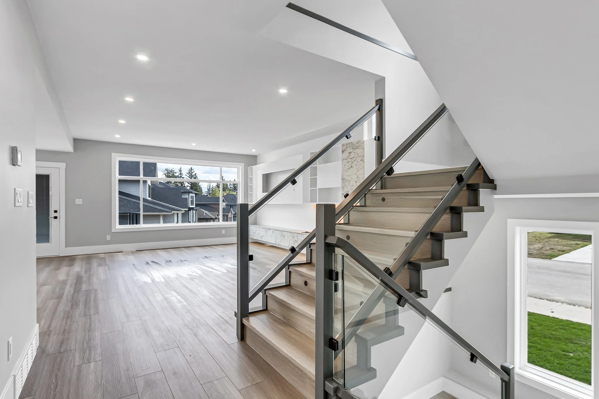 Modern living room with wood flooring, large window, staircase with glass and metal railing, and white walls.