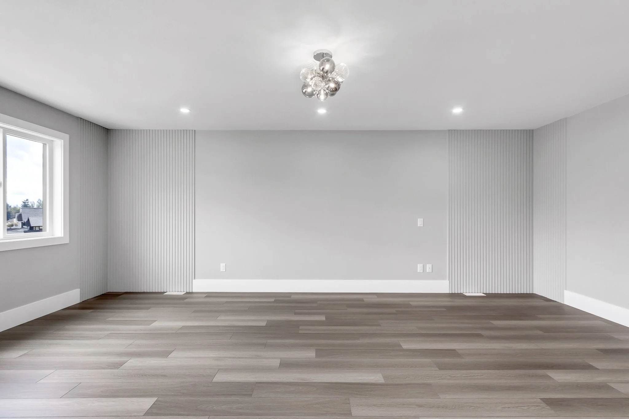 Empty living room with gray walls, white baseboards, wooden floor, window on left, modern ceiling light fixture.