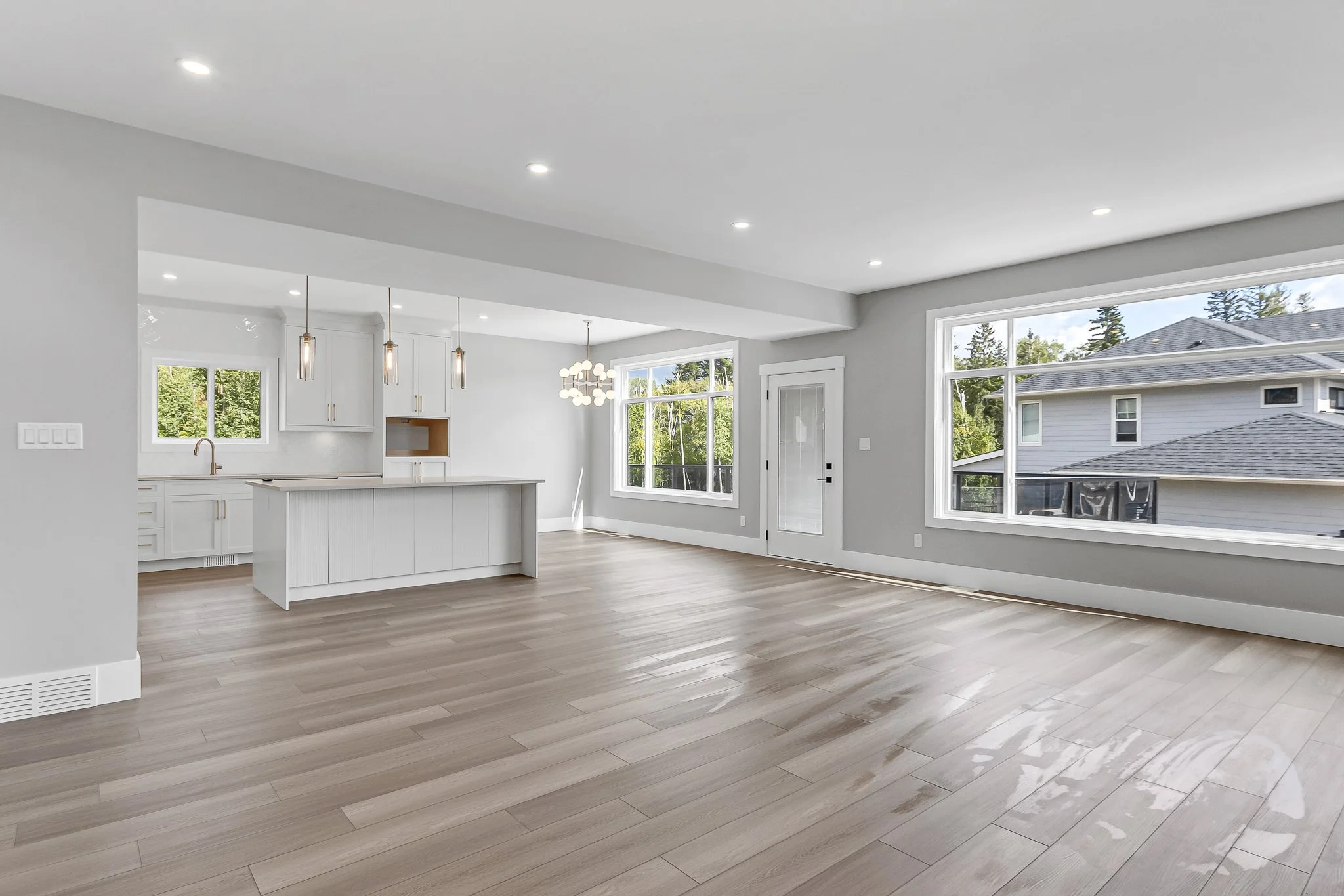 Empty modern living room and kitchen with large windows, light wood flooring, white walls, and natural light.