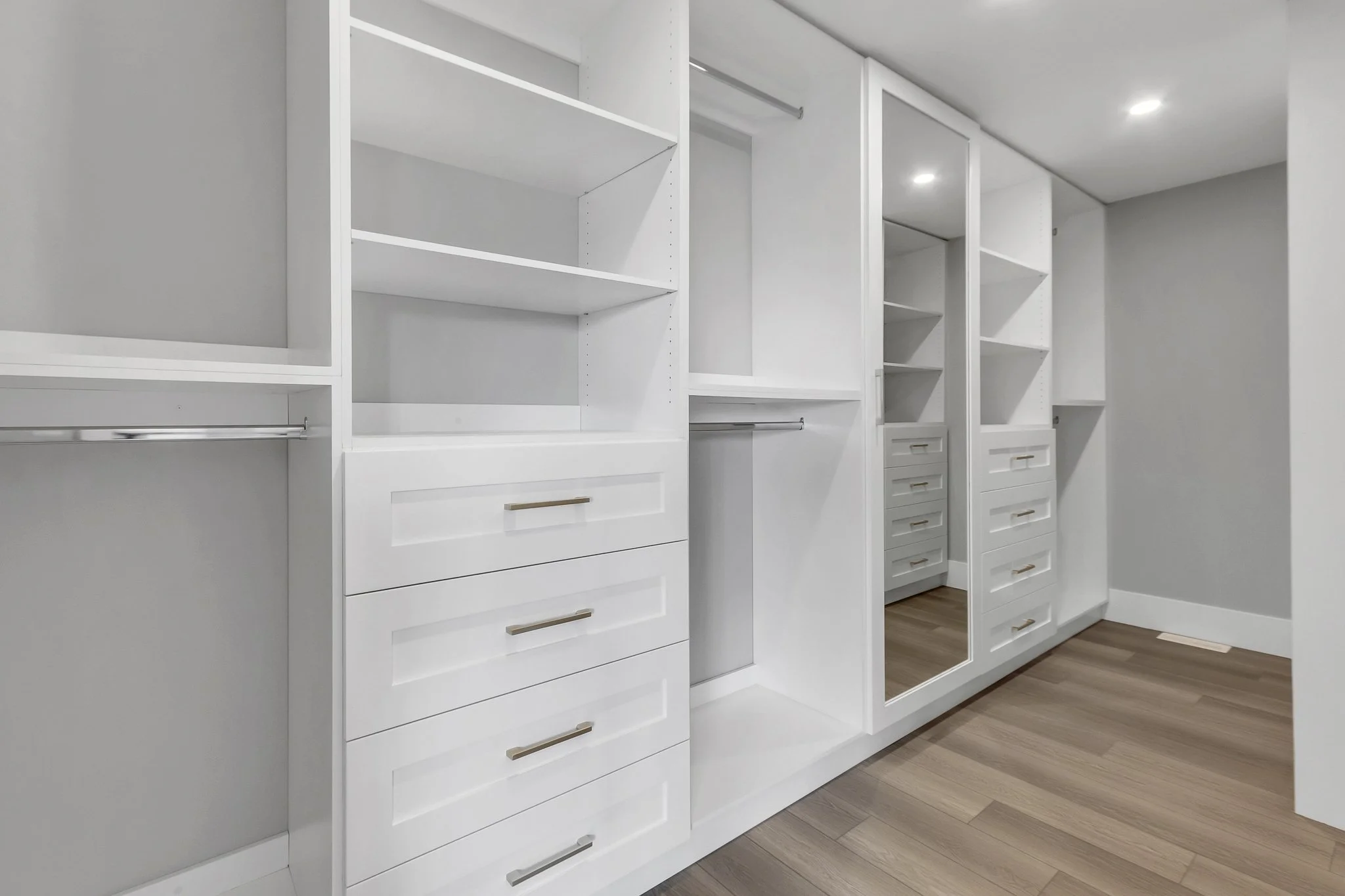 Empty white walk-in closet with built-in shelves, drawers, and hanging rods, hardwood flooring, and recessed lighting.