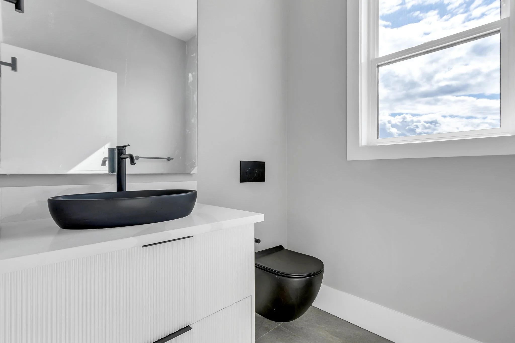 A minimalist bathroom with a black sink on a white vanity, a black wall-mounted toilet, a large window with blue sky and clouds, and gray flooring.
