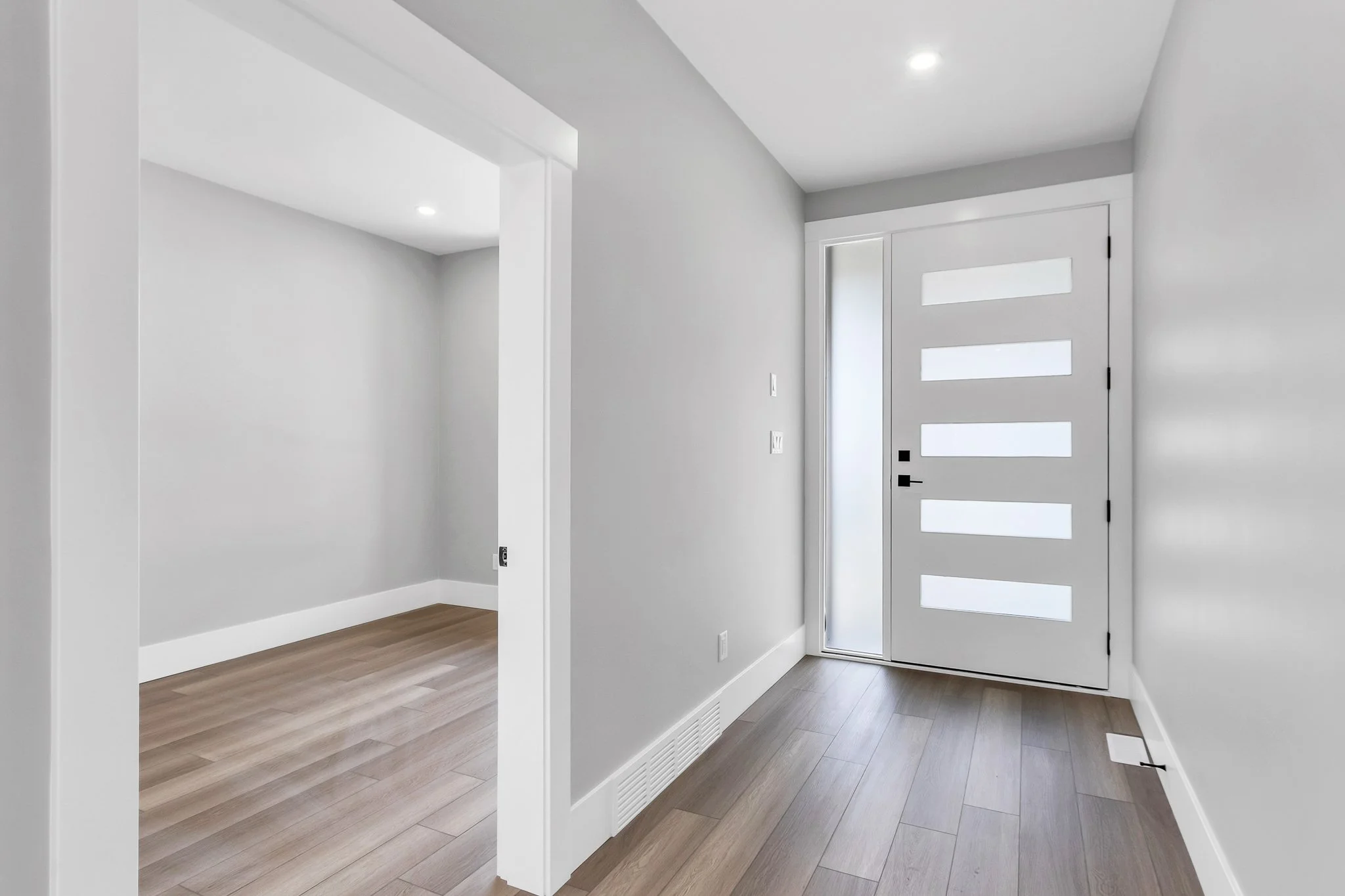 Modern, minimalistic entryway with white door featuring horizontal glass panels, light-colored wooden flooring, and gray walls.