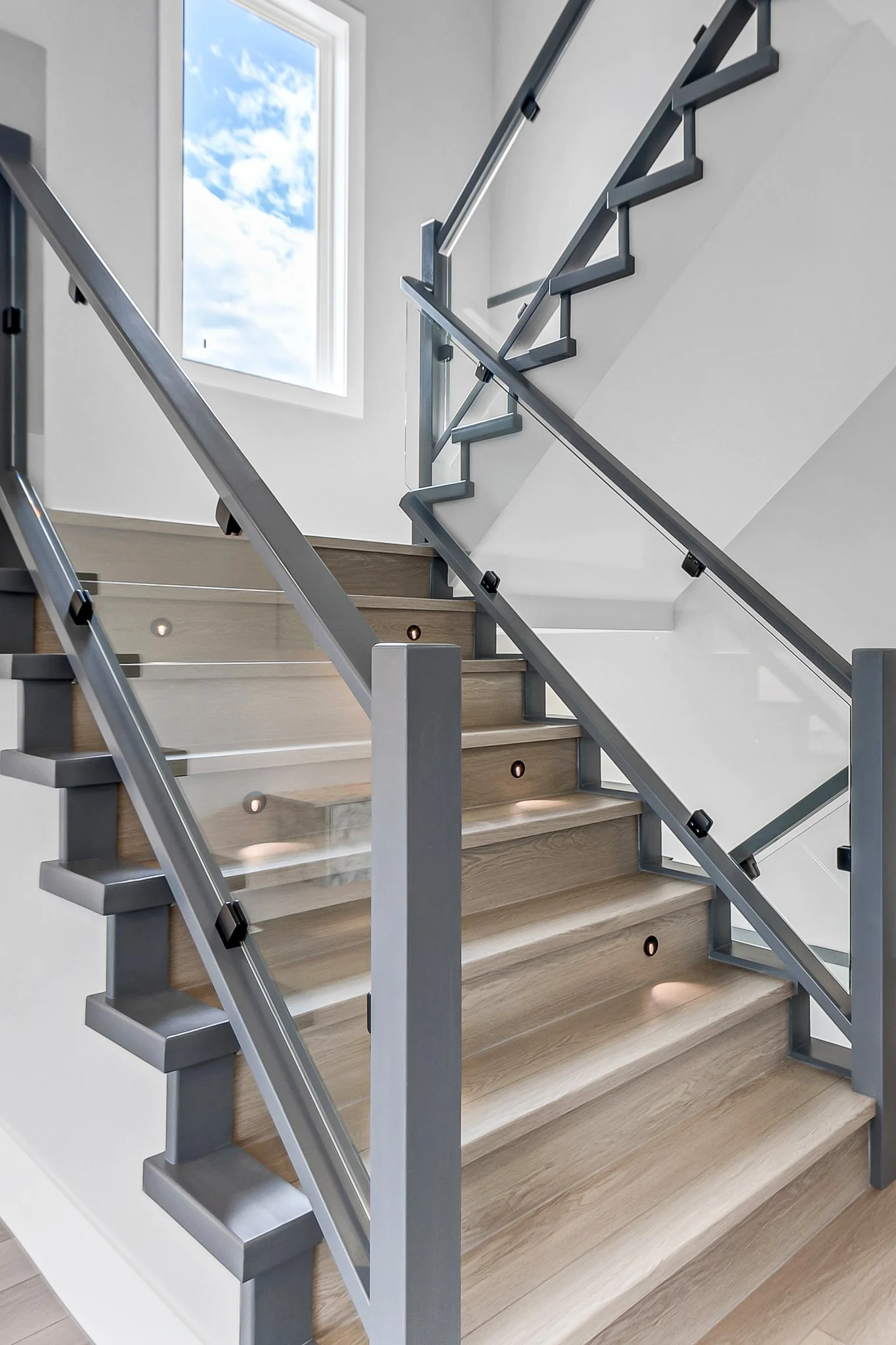 Modern staircase with wooden steps, metal handrails, glass panels, and recessed lighting near a window showing a partly cloudy sky.