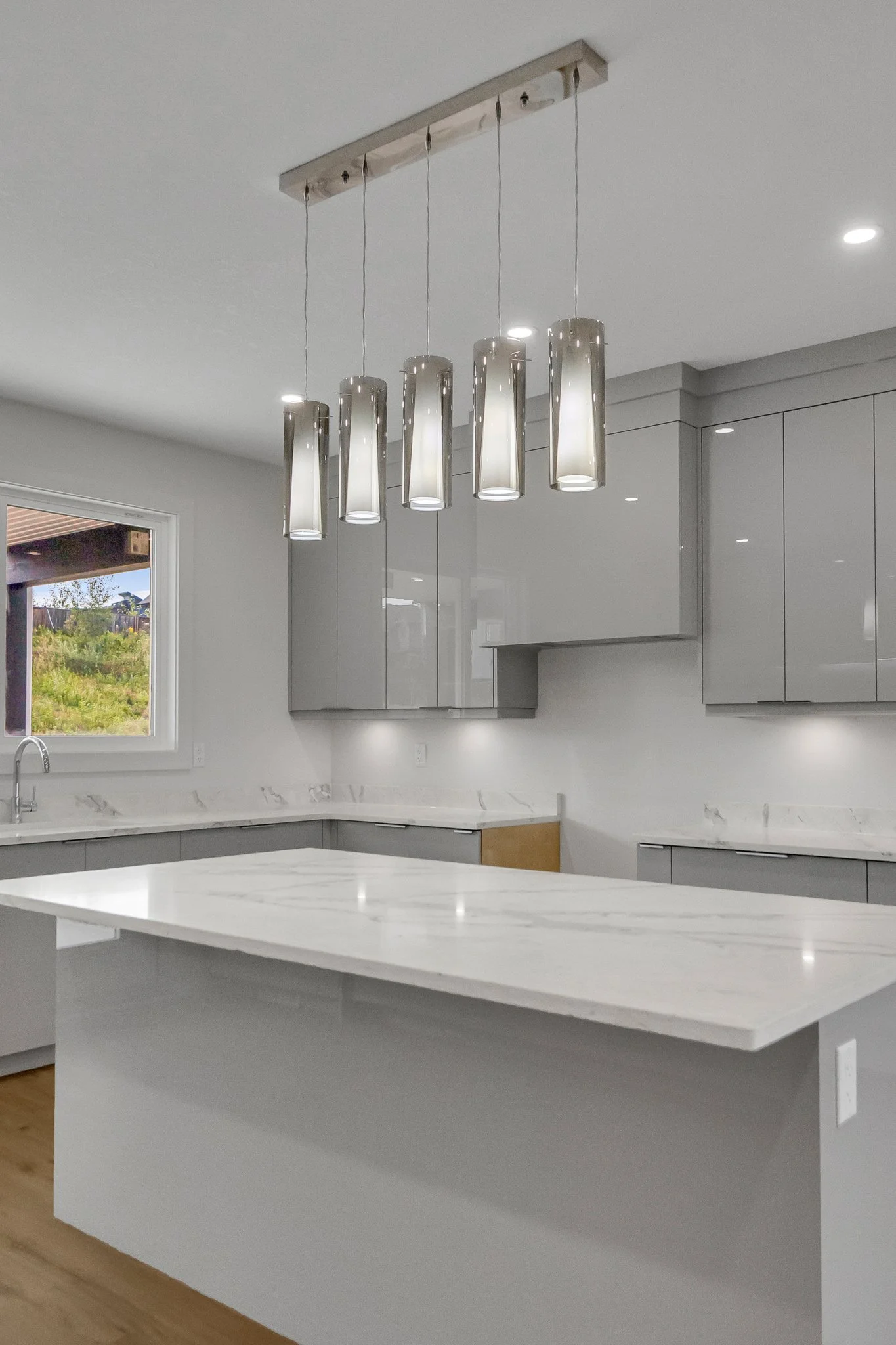 Modern kitchen with quartz countertops, gray cabinets, a window, and a rectangular pendant light fixture with six cylindrical glass lights.