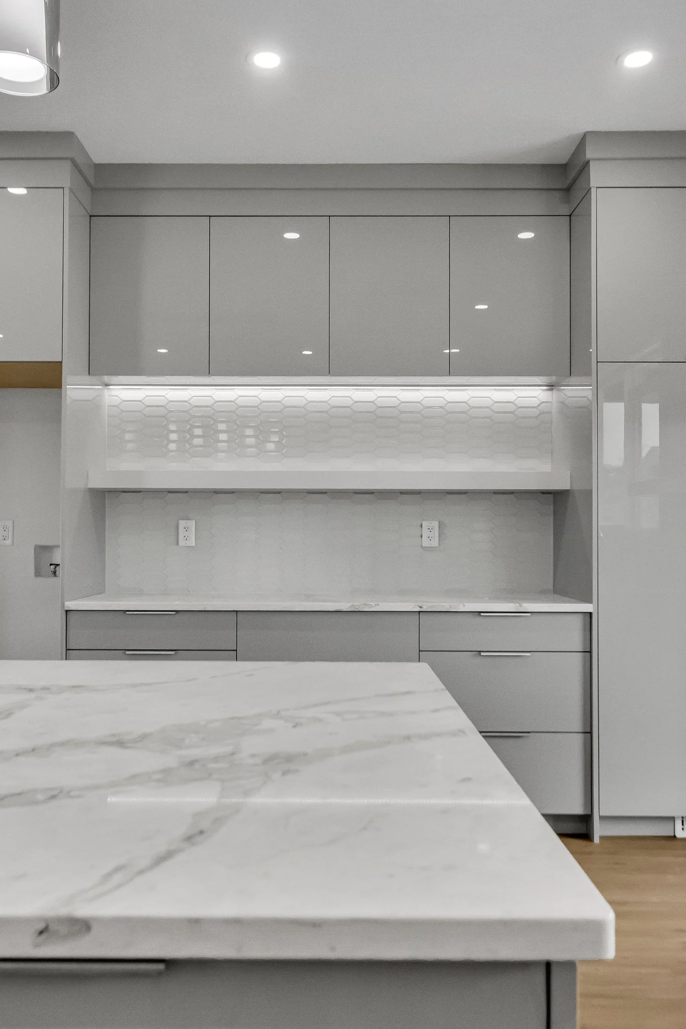 Modern kitchen with white and gray cabinets, marble countertop, and white subway tile backsplash with a honeycomb pattern, illuminated by recessed ceiling lights.