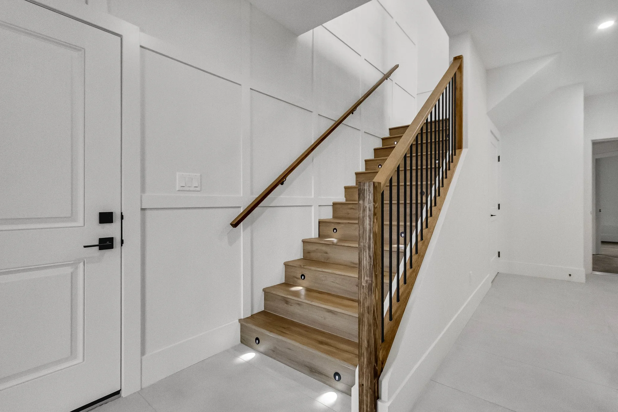 Interior view of a modern staircase with wooden steps and black metal railing, embedded with small round lights in the treads, next to a white paneled wall and a white door.