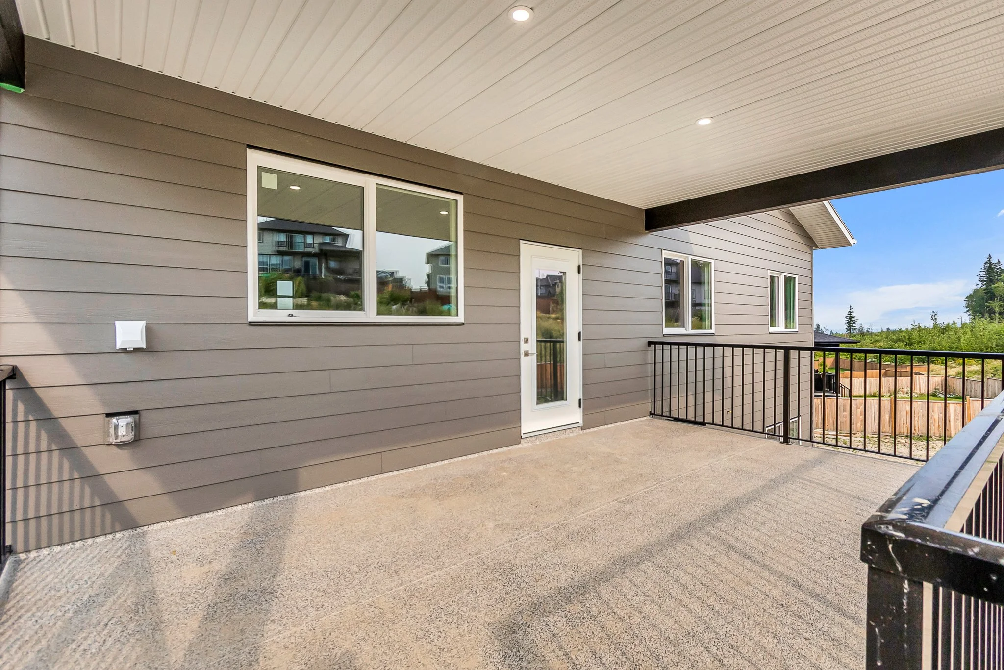 Balcony with concrete floor, gray siding walls, black metal railing, white-framed window and door, view of residential neighborhood and trees in the background, under a white wood-paneled ceiling with recessed lighting.