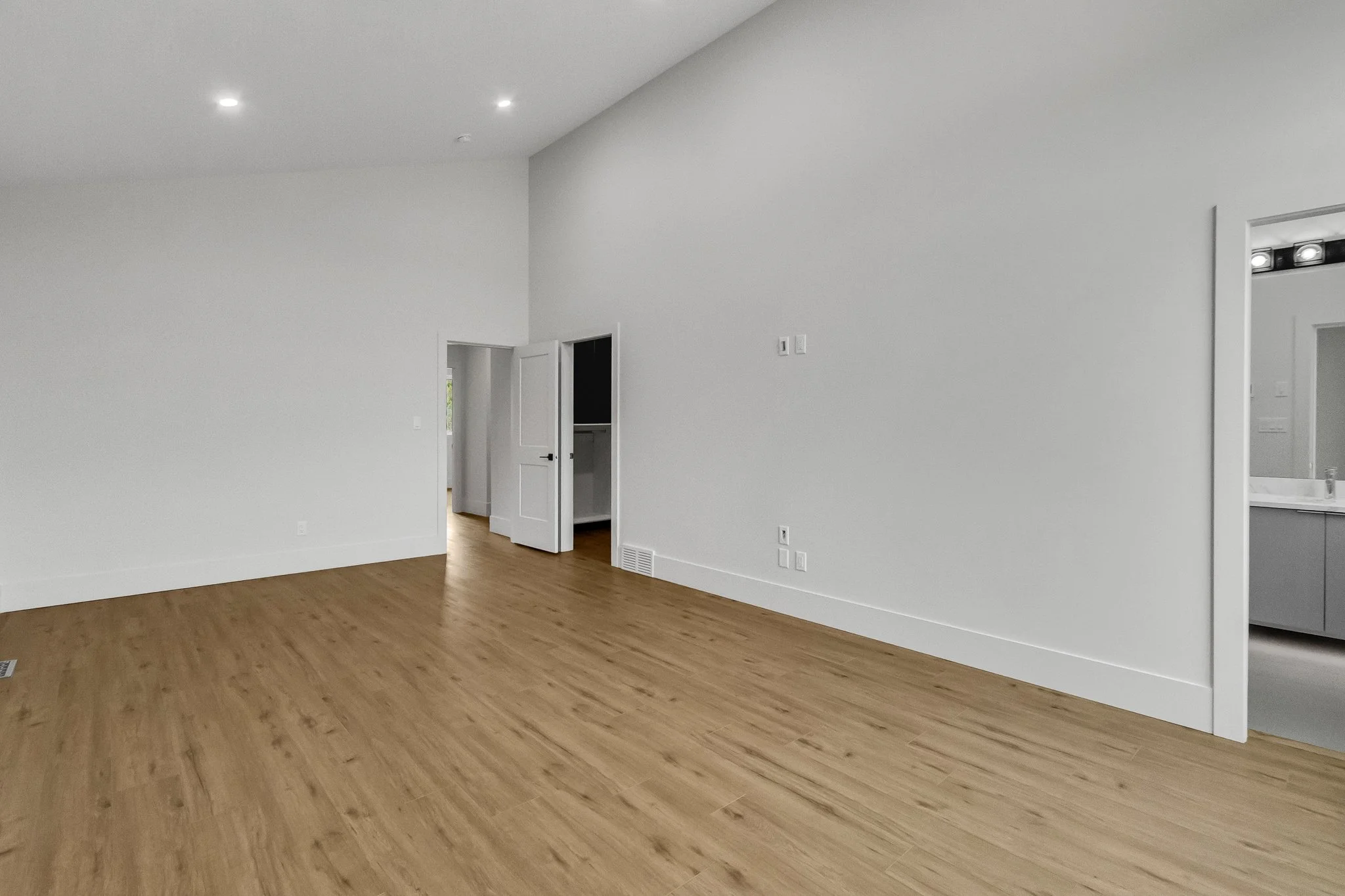 Empty room with white walls, wooden floor, and an open doorway leading to a hallway and a bathroom.