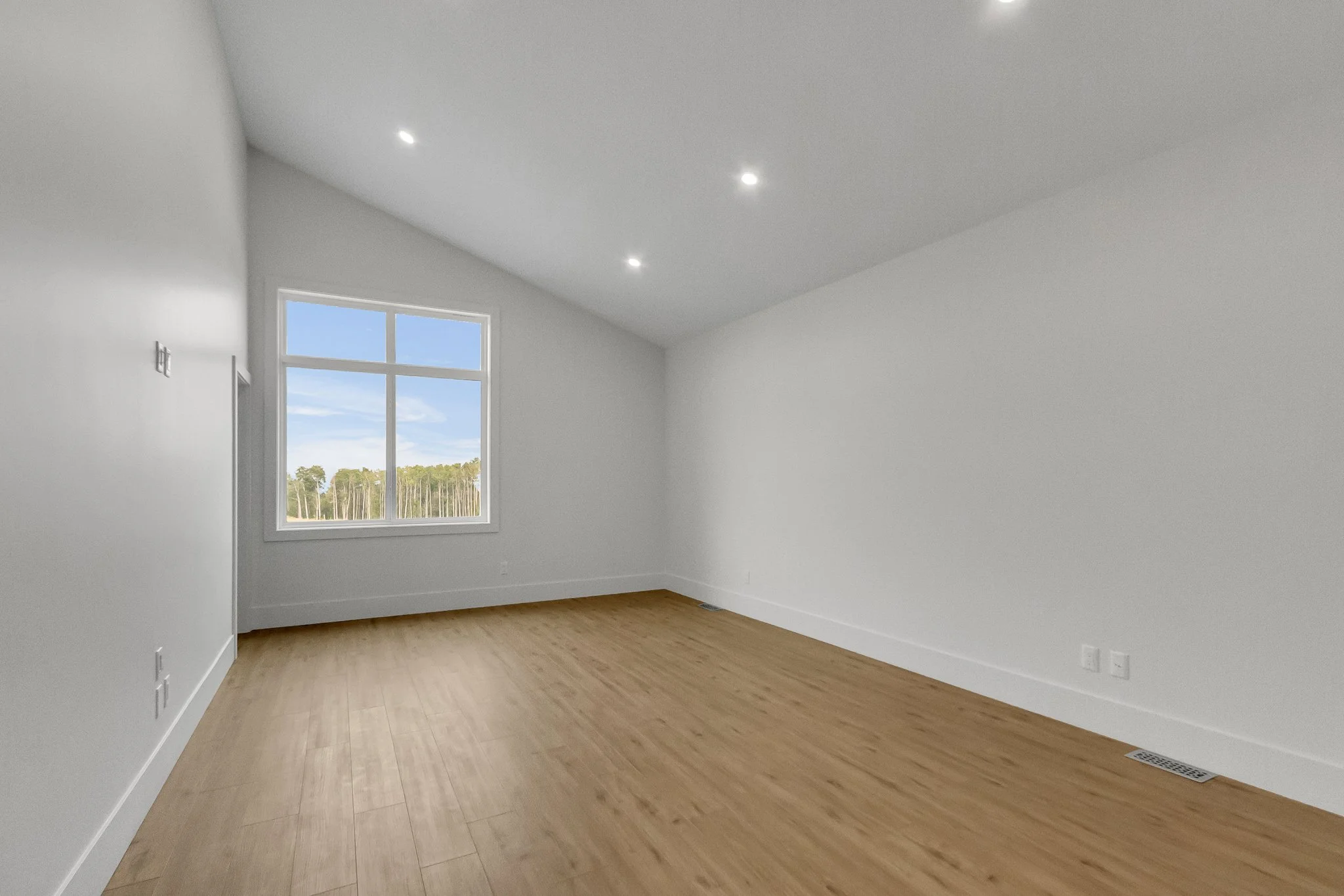 Empty room with white walls, hardwood floor, large window with view of trees, ceiling with recessed lighting.
