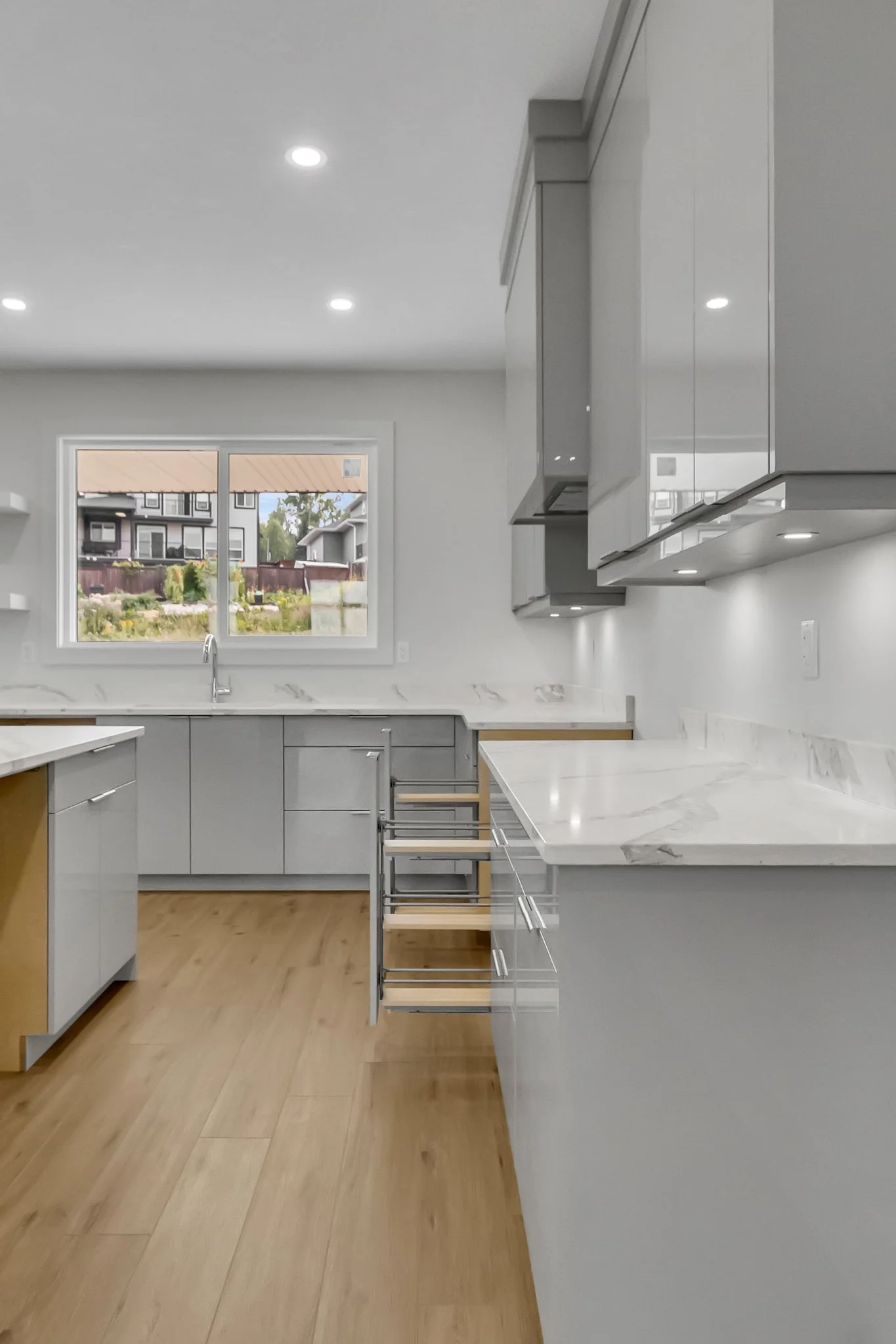 Modern kitchen with light gray cabinets, marble countertops, wooden floor, window showing residential neighborhood, under-cabinet lighting, and empty drawer slides.