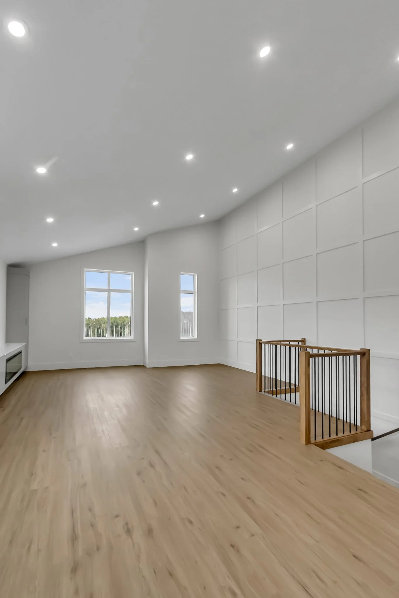 Empty modern room with wooden floor, white walls, ceiling lights, and two large windows showing trees outside, with a wooden staircase railing.