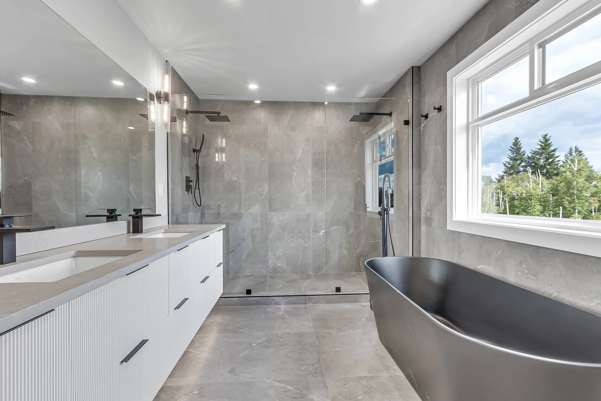 Modern bathroom with a double vanity, wall-mounted faucets, grey marble walls and floor, a black freestanding bathtub, and a large window showing trees outside.
