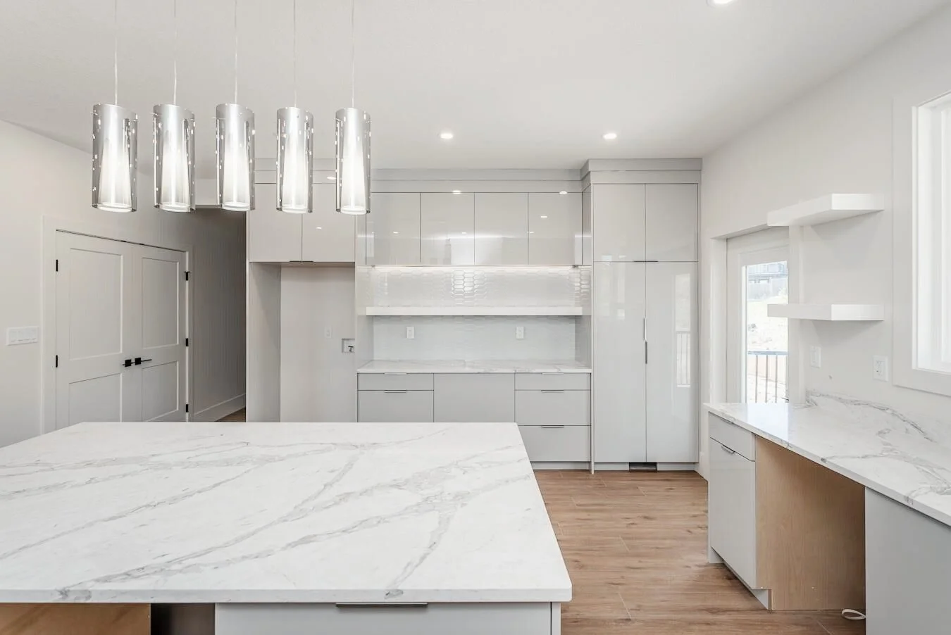 Modern white kitchen with marble countertops, pendant lights, and natural light from windows and glass doors.