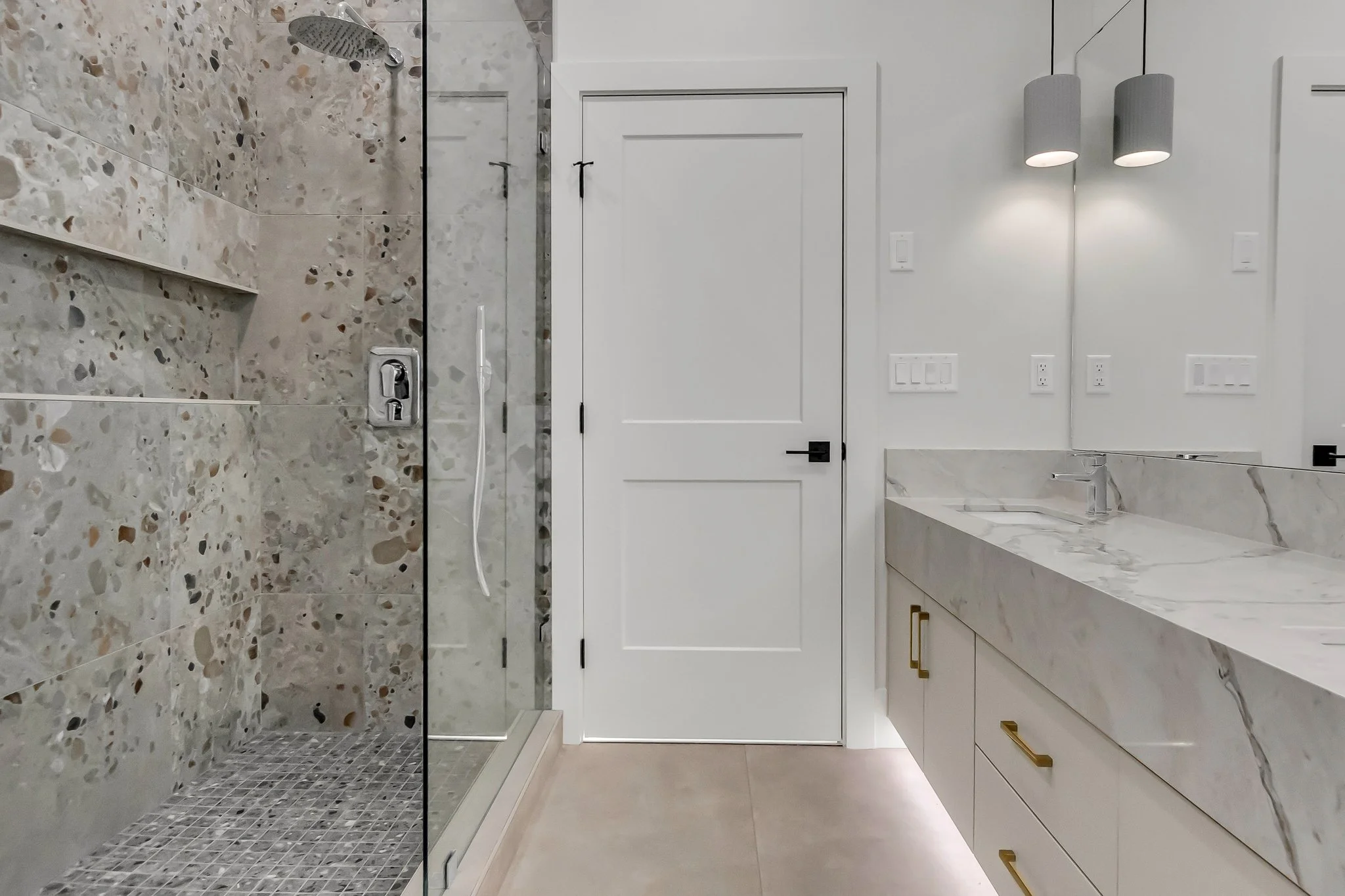 Modern bathroom with a glass-enclosed shower, marble vanity with gold handles, wall-mounted lights, and a white door.