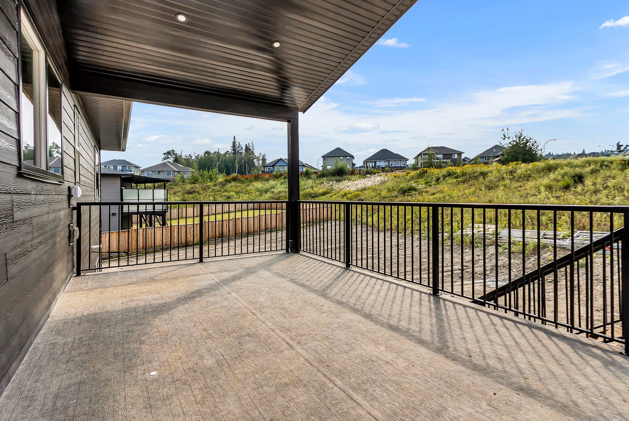 Empty patio with black metal railing overlooking a grassy hill with houses in the distance and partly cloudy sky.