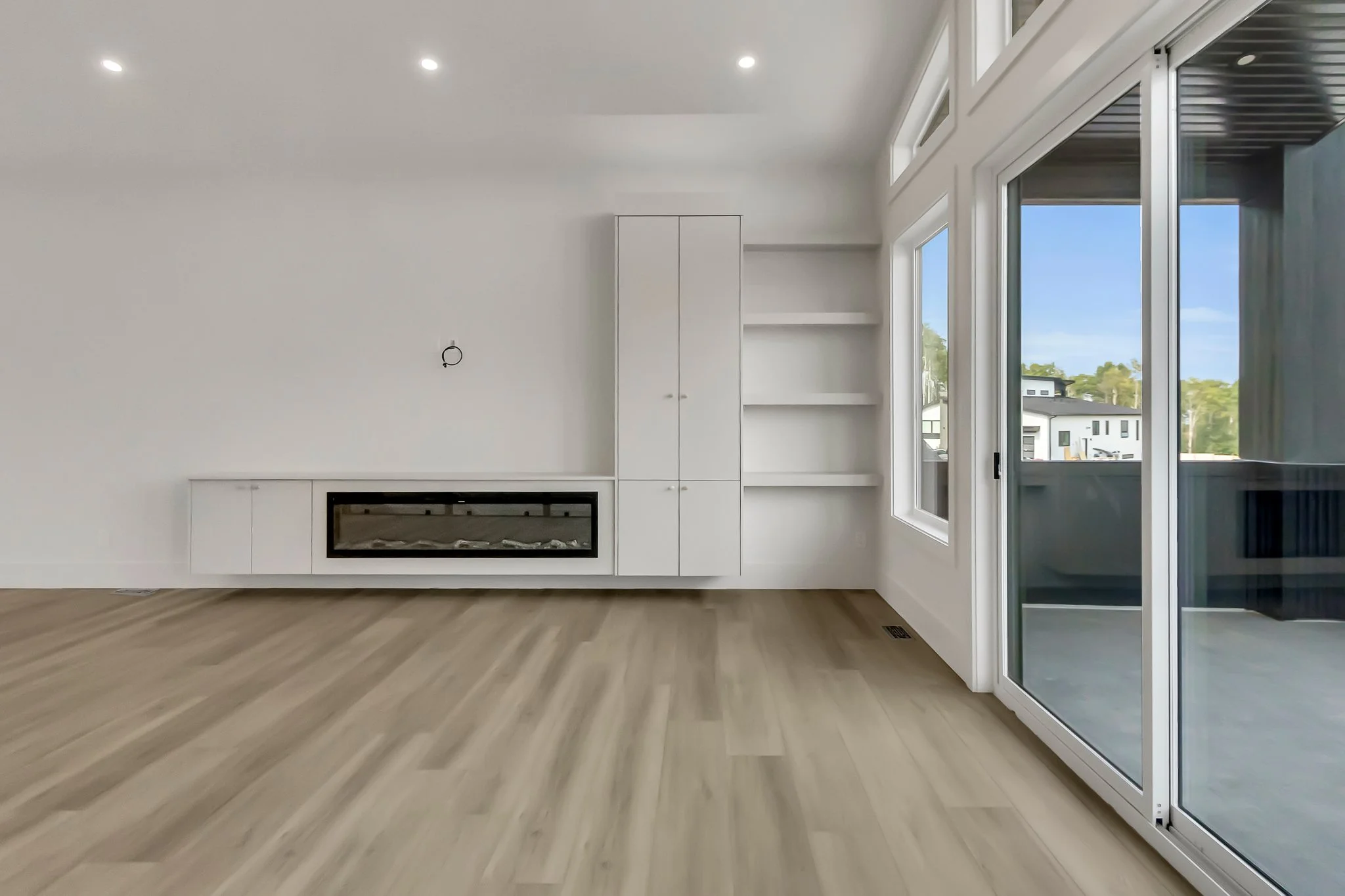Empty living room with white walls, a modern electric fireplace, built-in shelves, large glass sliding door, and light wood flooring.