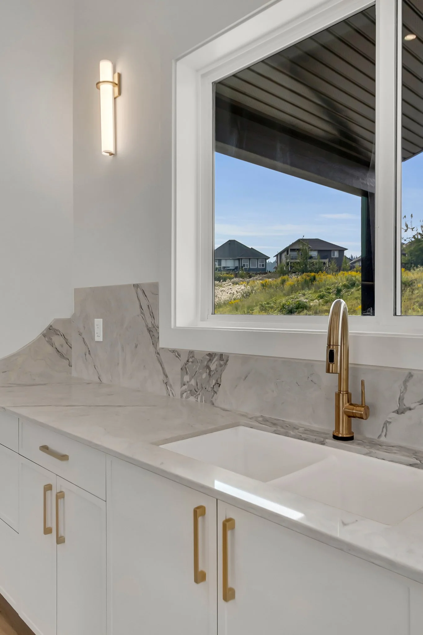 Kitchen sink with a gold faucet, white cabinets with gold handles, marble countertop and backsplash, window showing houses and green landscape outside, wall-mounted light fixture.