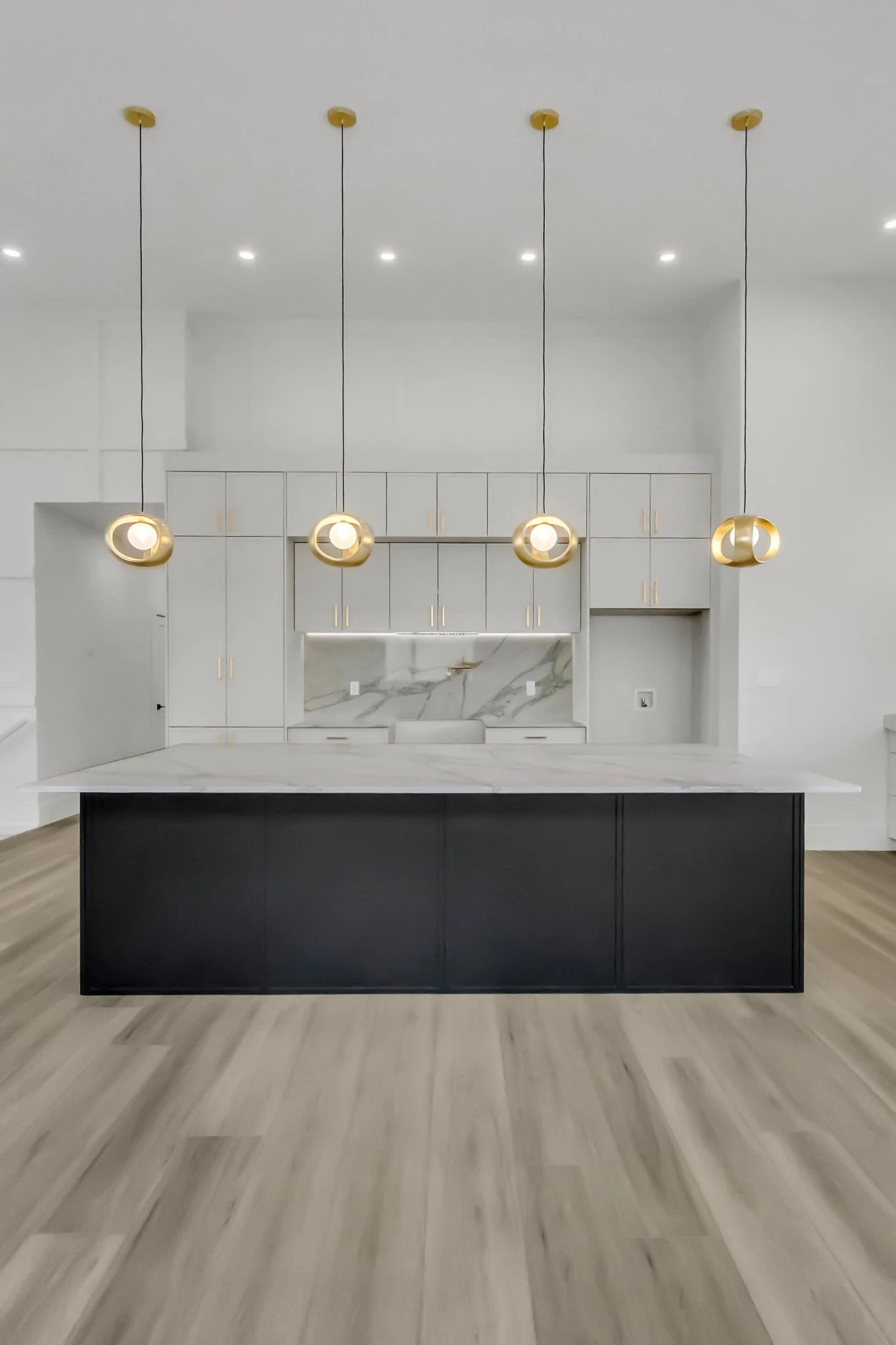 Modern kitchen with a large island, white marble countertops, gold pendant lights, white cabinets with gold handles, and light wood floors.