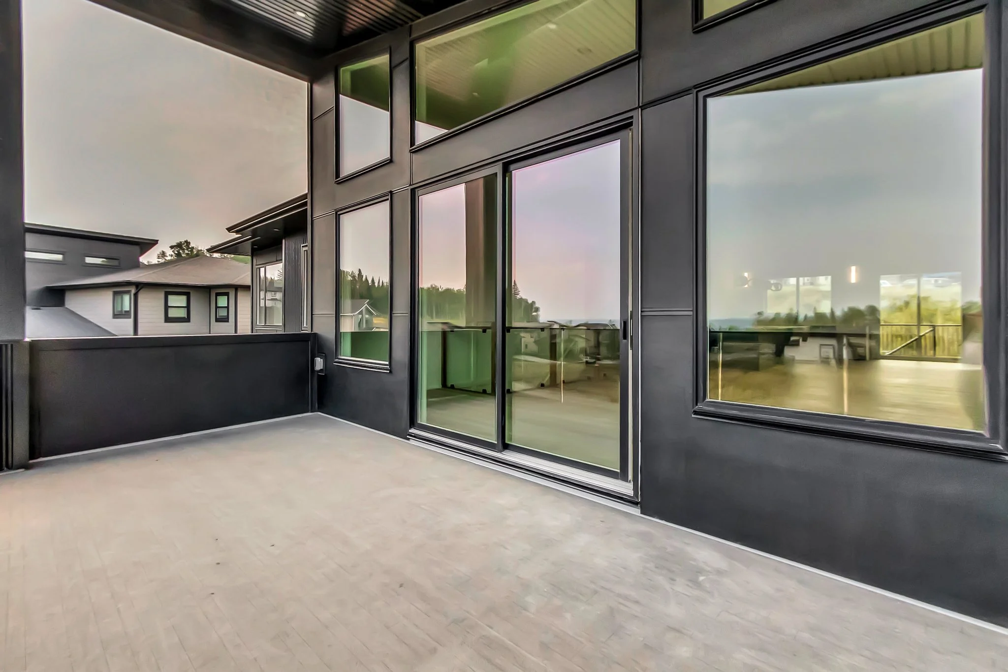 Empty balcony with glass sliding door and black exterior walls, overlooking neighboring houses and a green landscape.