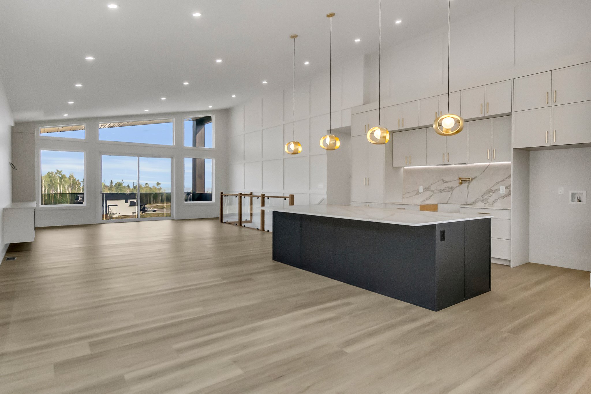 Modern kitchen and living area with large windows, light wood flooring, white cabinets, black kitchen island, and gold pendant lights.