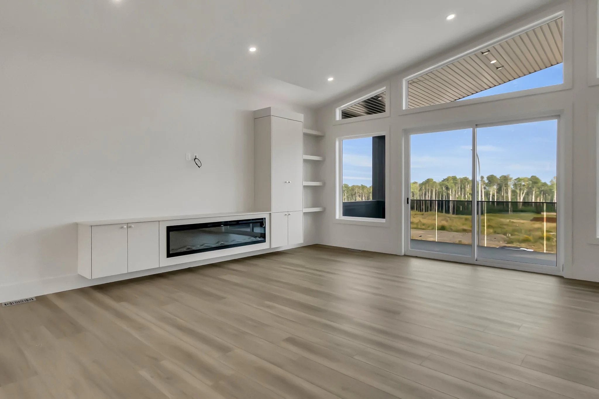Empty room with large windows and sliding glass door, white walls, light wood flooring, built-in white shelves and a modern electric fireplace.