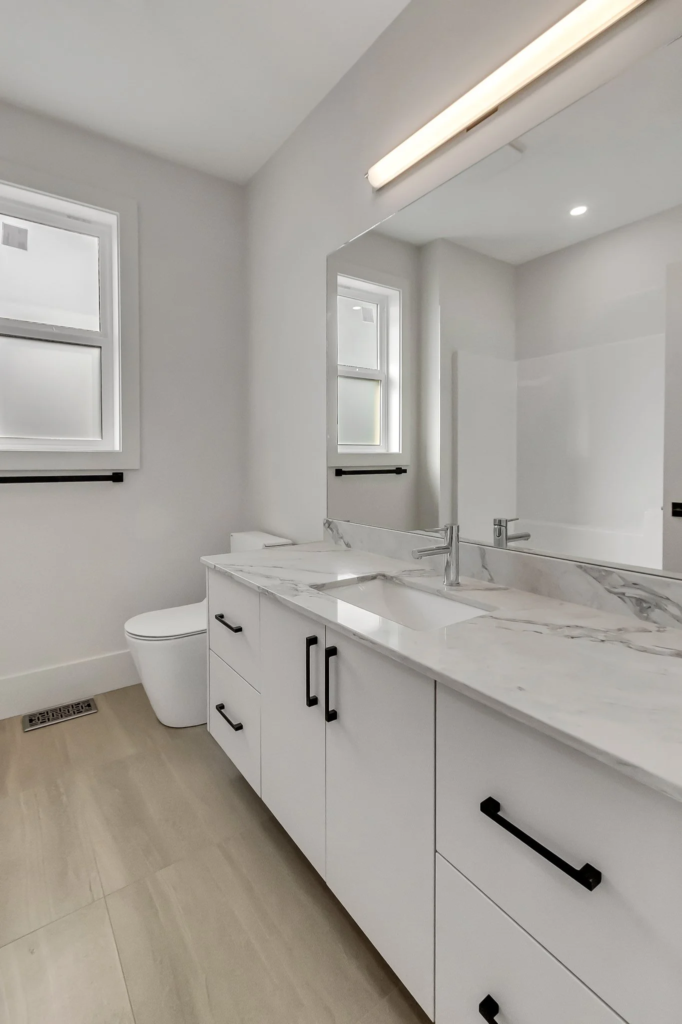Modern bathroom with white vanity and marble countertop, large mirror, window, and beige tiled floor.