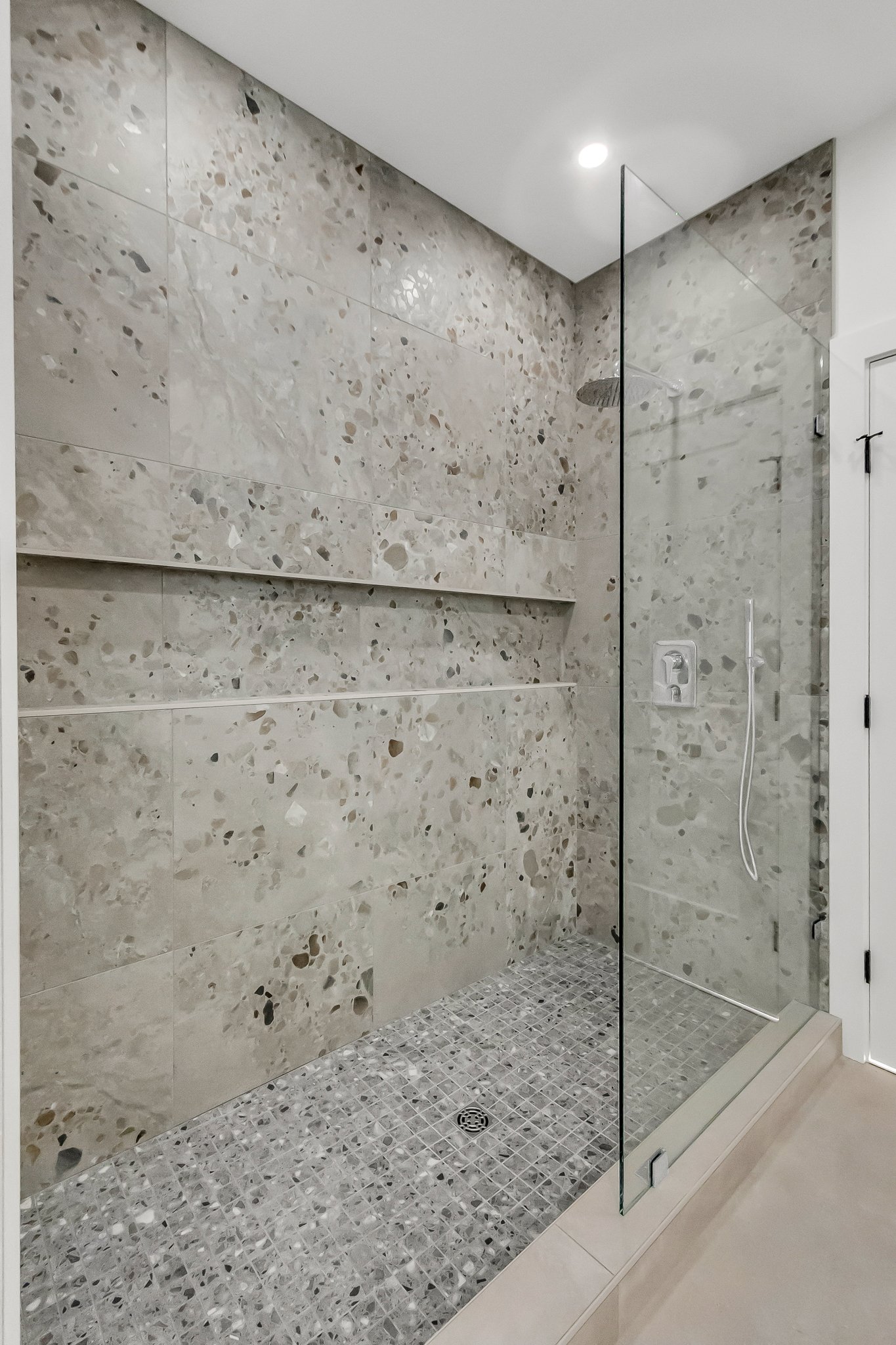 Modern walk-in shower with beige stone tiles on the walls, small gray mosaic tiles on the floor, a built-in bench, a ceiling rain shower head, and a glass panel door.