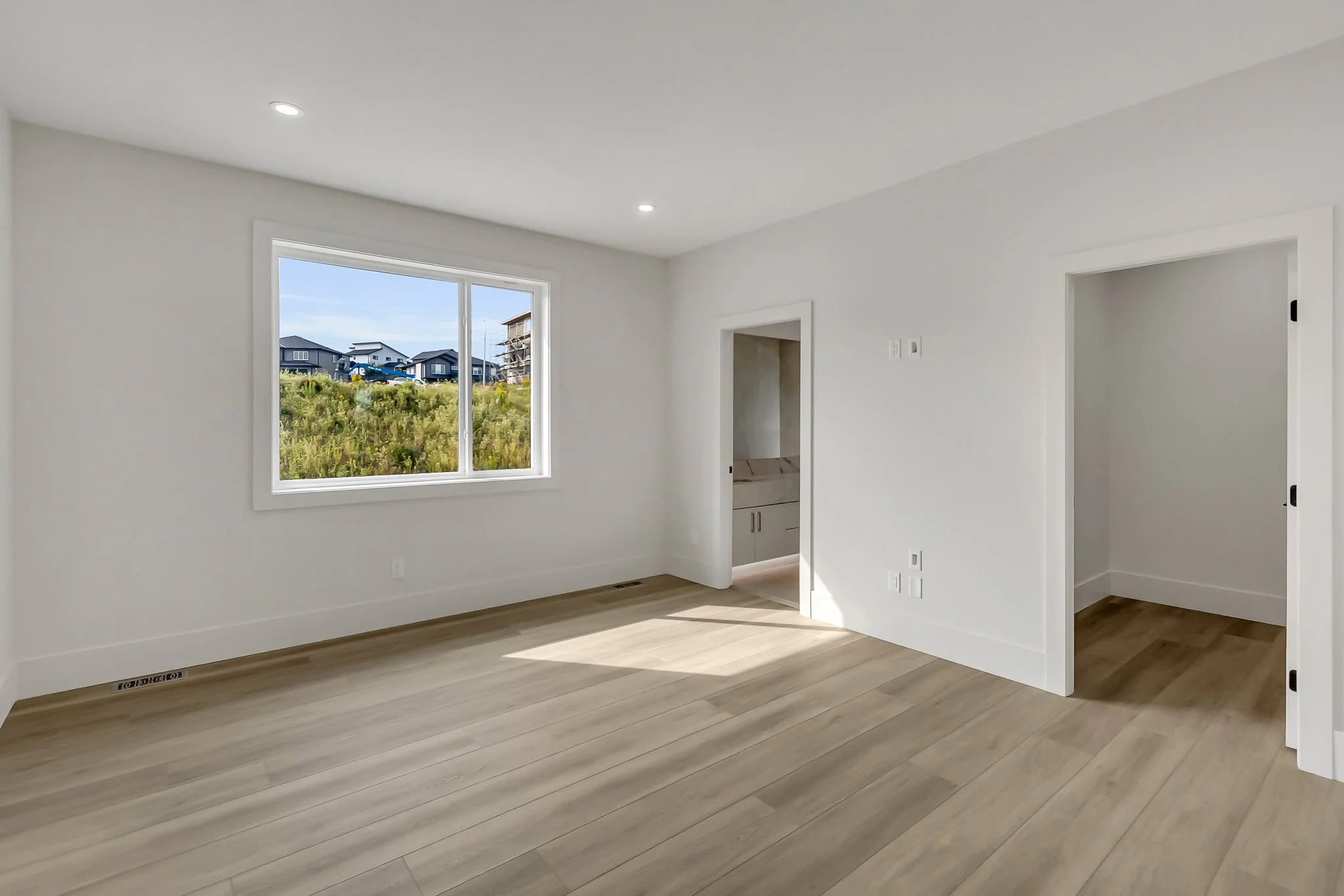 Empty room with white walls, hardwood floor, a window showing an outdoor view of houses, and a small open closet.