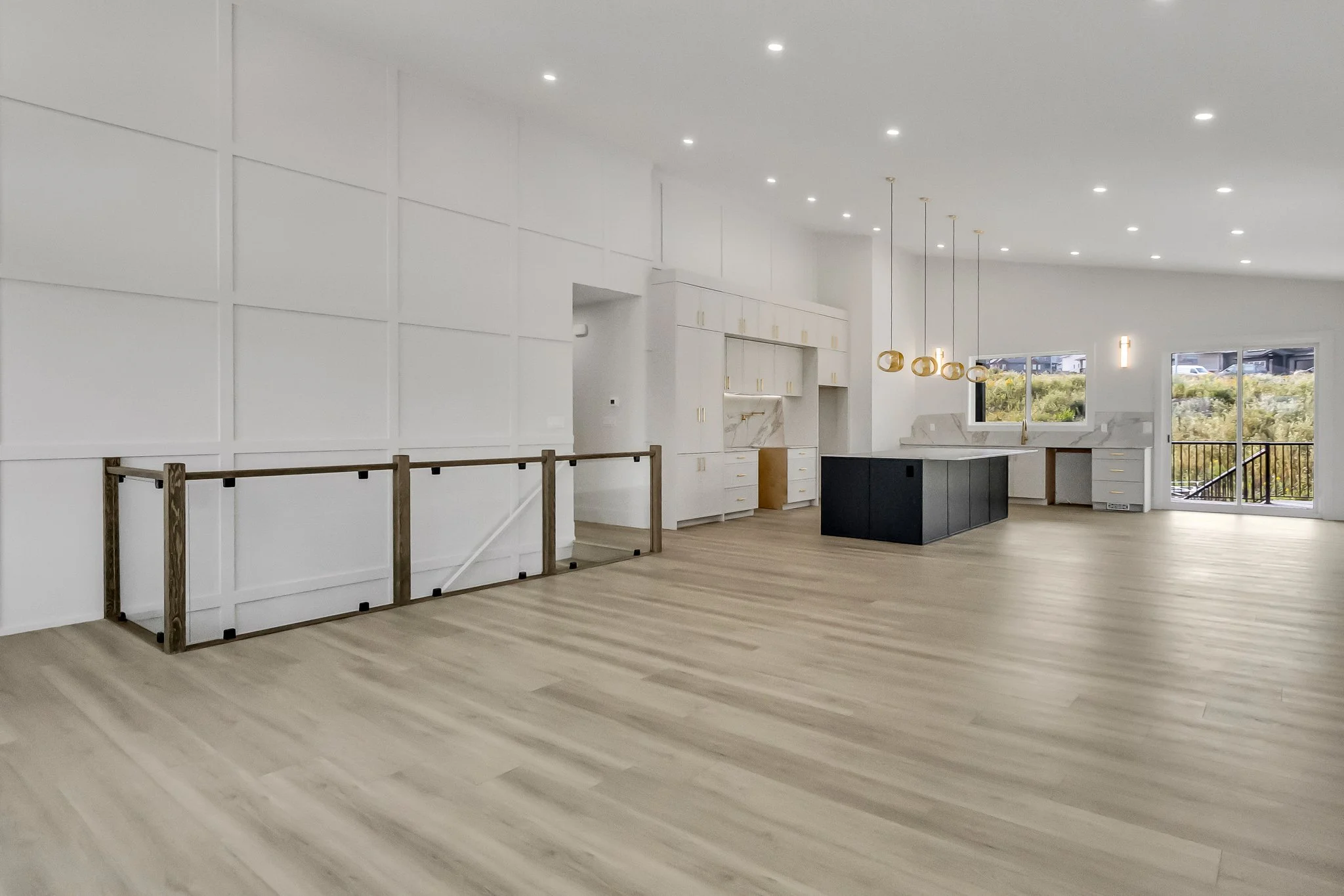 Empty modern open-concept kitchen and living space with light wood flooring, white walls, and large windows. Features a black kitchen island with hanging gold pendant lights.