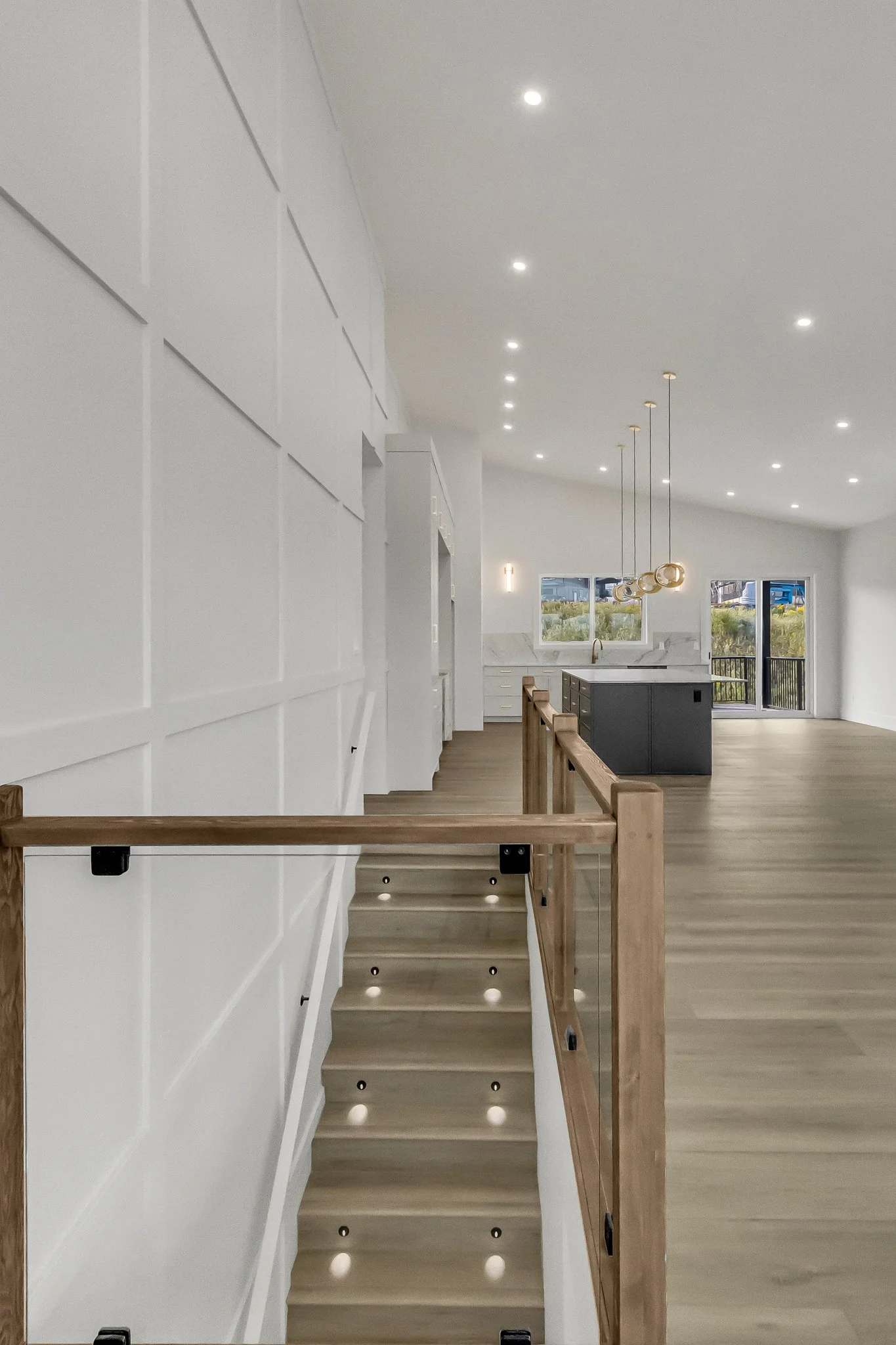 Interior of a modern, open-concept kitchen and living space with white walls, wooden flooring, and a kitchen island with a dark base; large windows and a balcony door on the far side.