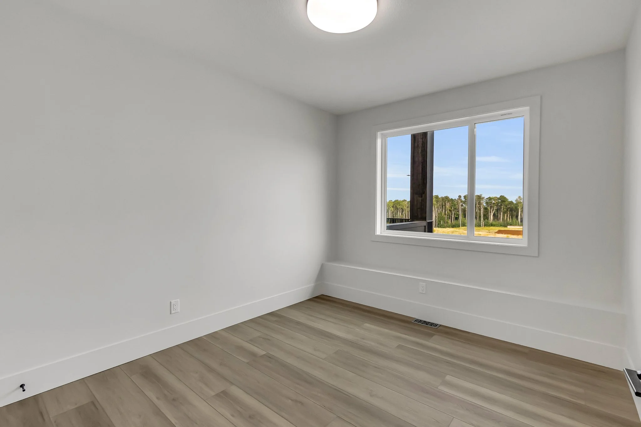 Empty room with white walls, wood flooring, a window showing outdoors, and a ceiling light fixture.