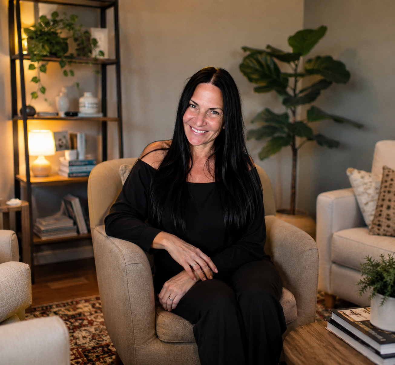 A woman with long black hair sitting on a beige armchair, smiling, in a cozy living room with a bookshelf, lamp, and plants.