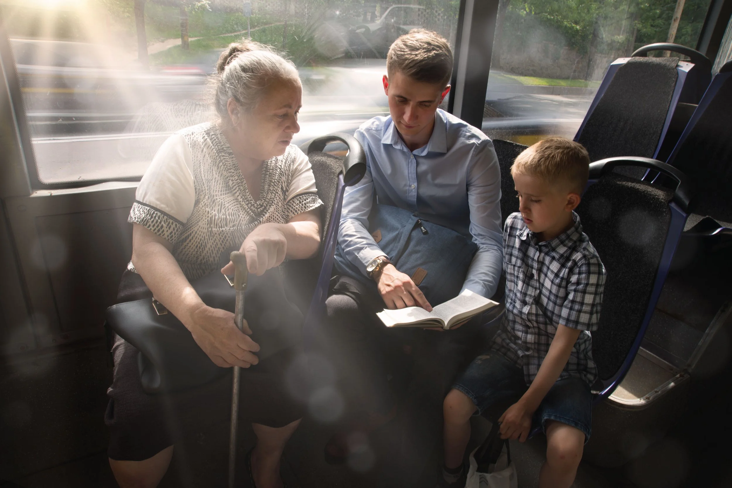 An elderly woman with gray hair and a young man with short brown hair sitting on a bus looking at a book together. A young boy with short light hair is sitting next to them. The woman is in a wheelchair, and the sunlight is coming through the bus window.