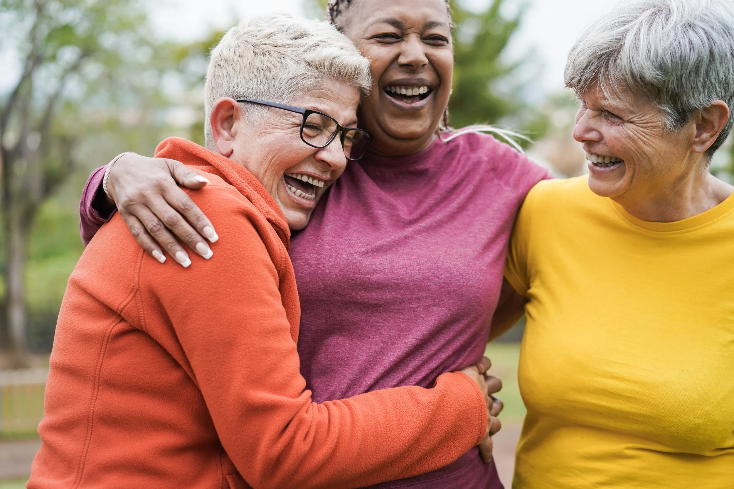Three women sharing a joyful group hug outdoors, smiling and laughing in a park.