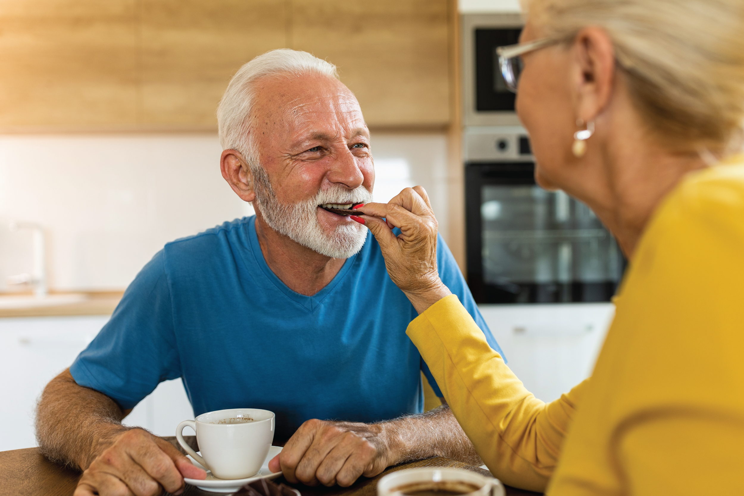 An elderly man with a white beard smiling and having his wife feed a piece of chocolate to him at the kitchen table. The man is wearing a blue shirt and there are cups of coffee on the table.
