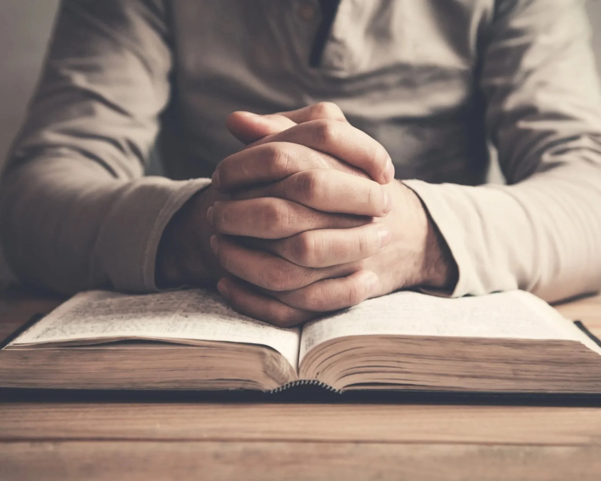 A person sitting at a table with hands clasped on top of an open bible.