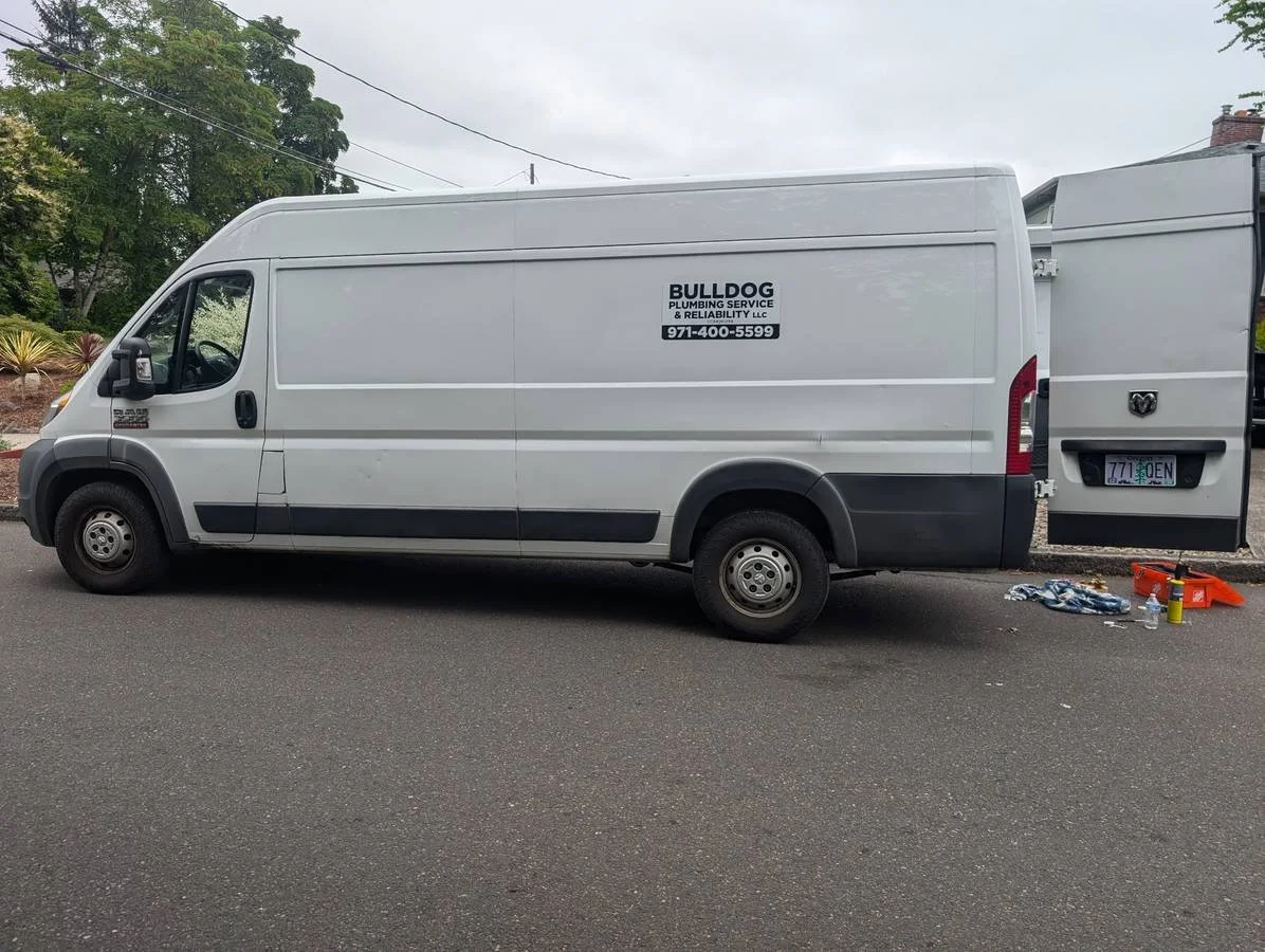 A white service van parked on the street with its rear door open, displaying signage for Bulldog Plumbing Service & Reliability LLC, with a phone number. Tools and supplies are on the ground near the open door.