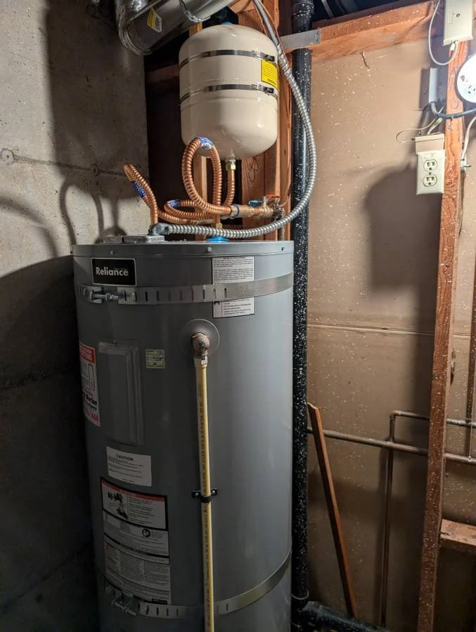 A Reliance water heater installed in a utility room, with a pressure tank, copper and flexible pipes connected, and an electrical outlet nearby.