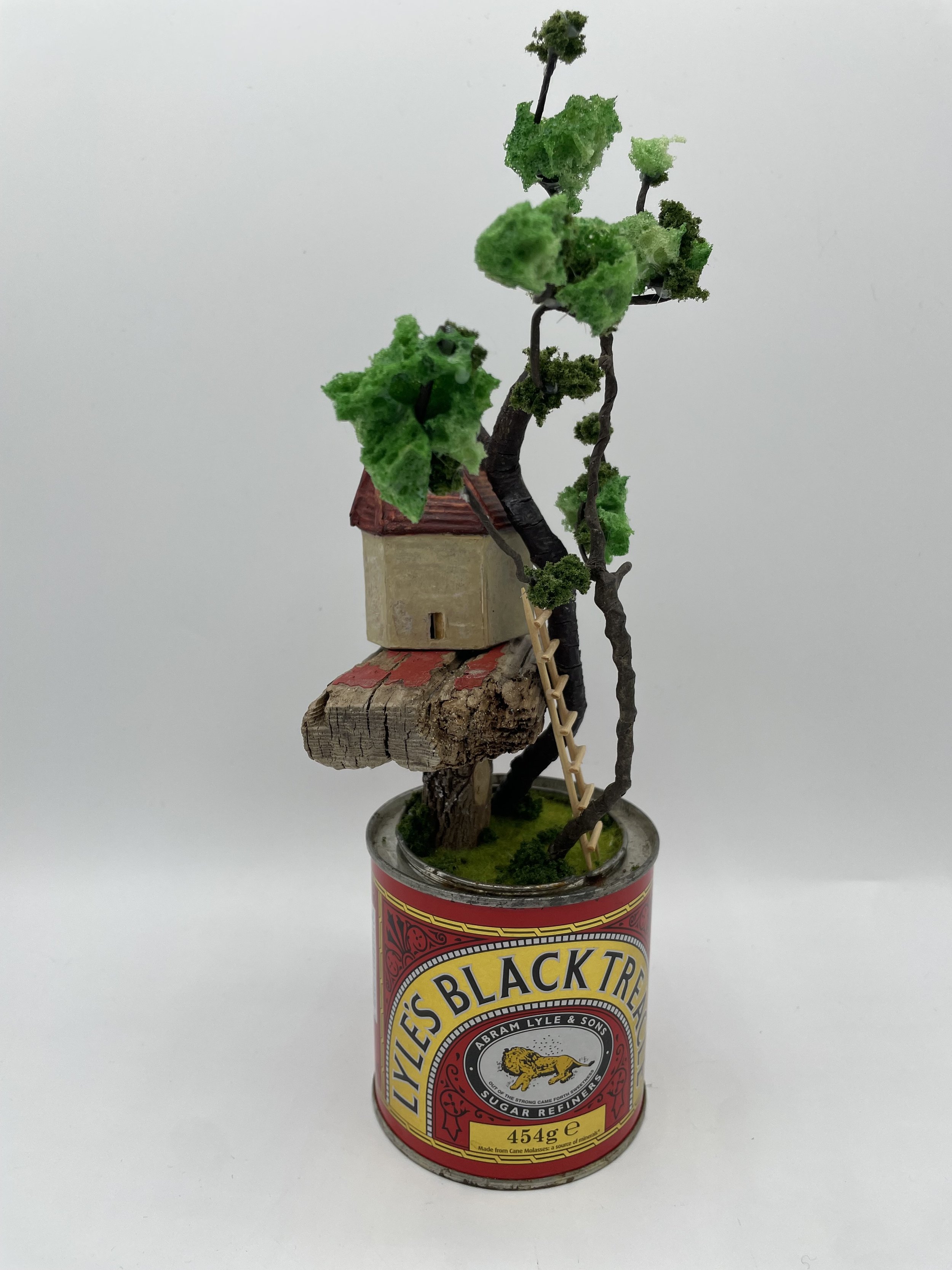 Miniature model of a treehouse with green foliage, a small ladder, and a tiny house on a branch, placed on top of a can of Lyle's Black Treacle.